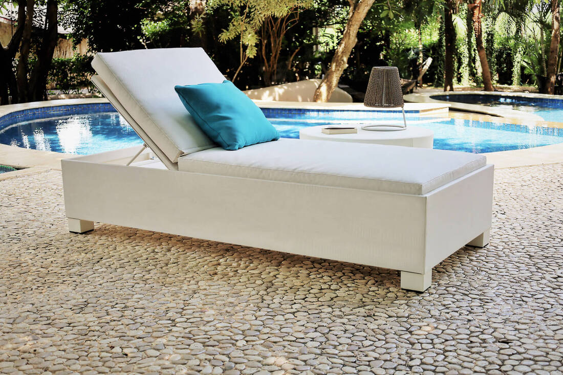 White upholstered sunbed with blue decorative cushion, side table with garden lamp in a pool terrace with white pebbles floor in a tropical garden in Sotogrande, cadiz, Spain